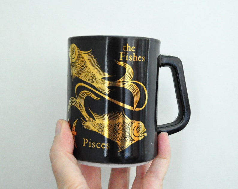 以下が含まれることがあります： 金色の魚2匹と「the Fishes Pisces」の文字が描かれた黒いセラミック製のマグカップです。