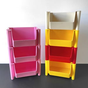May include: Two sets of colorful, stackable storage bins. One set is pink, the other is yellow, red, and off-white. Each bin has a rectangular shape with a cutout front for easy access. The bins are made of plastic and are designed for organizing items.