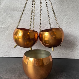 May include: Three polished copper-toned bowls with a smooth, reflective finish. Two of the bowls are suspended by thin brass-colored metal chains, each featuring three small, pointed feet at the base. One bowl rests below them on a flat surface.