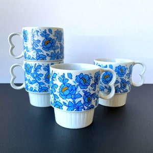May include: Set of four white ceramic mugs with blue and yellow floral pattern. The mugs have heart-shaped handles and stack on top of each other.