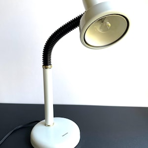May include: A white desk lamp with a flexible black neck and a round base. The lamp has a white shade and a visible lightbulb. The base has a small, gold-colored ring and a black power cord.