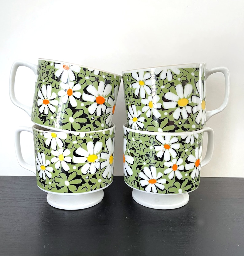 May include: Set of four white ceramic mugs with a green and black daisy pattern. The mugs have a footed base.