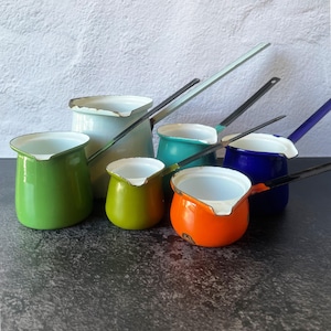 May include: A collection of six enameled metal pots with long handles, featuring a variety of vibrant colors including lime green, white, teal, mustard yellow, orange, and deep blue. Each pot has a white interior and a small pouring spout.