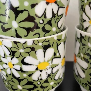 May include: Set of four white ceramic mugs with a green and black daisy pattern. The daisies have white petals, black centers, and yellow and orange centers.