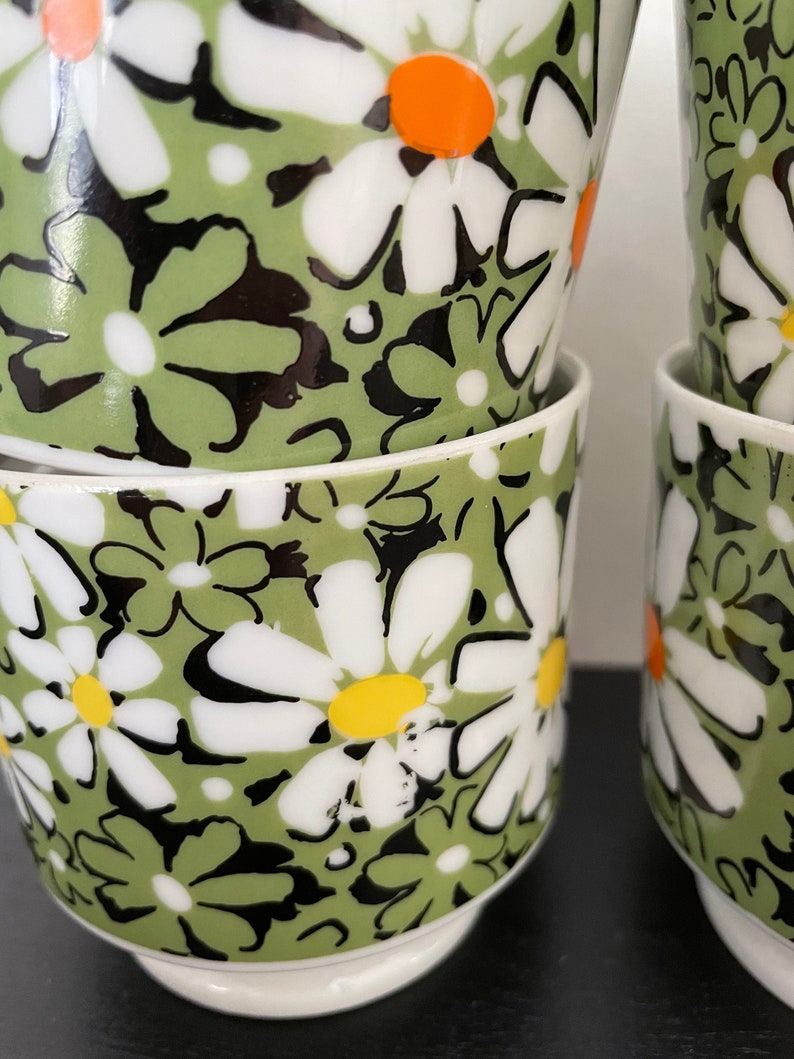 May include: Set of four white ceramic mugs with a green and black daisy pattern. The daisies have white petals, black centers, and yellow and orange centers.