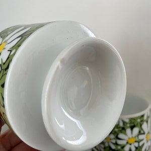 May include: White ceramic teacup with a green and white daisy pattern. The cup is shown from the bottom, revealing the white glazed interior.