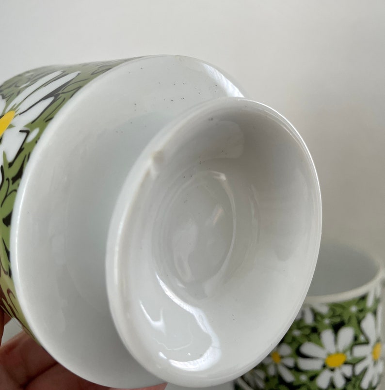 May include: White ceramic teacup with a green and white daisy pattern. The cup is shown from the bottom, revealing the white glazed interior.