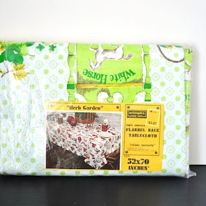 May include: A plastic-wrapped package containing a green and white flannel tablecloth with a floral pattern. The tablecloth is labeled "Herb Garden" and measures 52x70 inches.