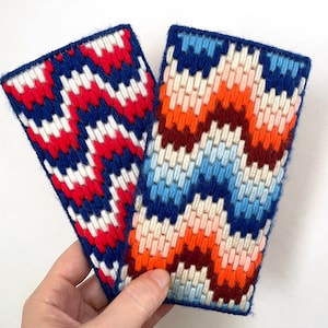 May include: Two colorful needlepoint wall hangings with geometric patterns. One is red, white, and blue, and the other is orange, blue, white, and brown.