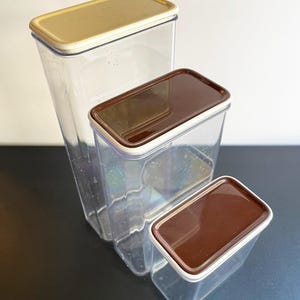 May include: Three clear plastic containers with lids. The largest container has a gold lid, the middle container has a brown lid, and the smallest container has a dark brown lid.