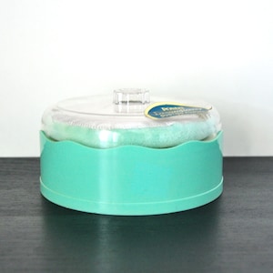 May include: A turquoise plastic container with a clear lid and a white puff inside. The container has a scalloped edge and the lid has a small knob on top. The label on the container reads "Powder Puffs Extra Soft"