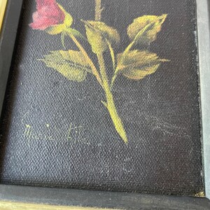 May include: A framed painting of a rose with a red bud, green stem, and yellow-green leaves. The artwork is set against a black background and framed with a gold and black frame. The artist's signature is visible.
