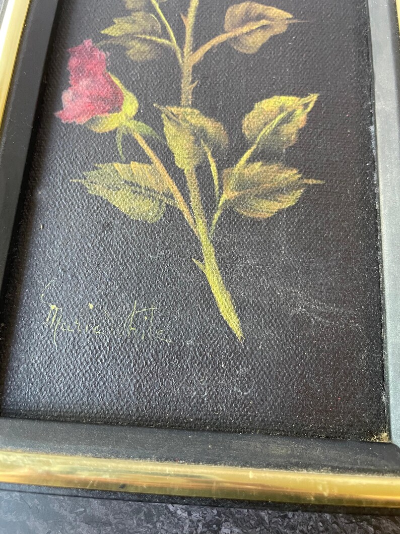 May include: A framed painting of a rose with a red bud, green stem, and yellow-green leaves. The artwork is set against a black background and framed with a gold and black frame. The artist's signature is visible.