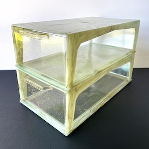 May include: Two-tiered, clear glass storage unit with a pale green frame. The unit has a rectangular shape with a flat top and two open compartments. Each compartment has a small, rectangular handle. The glass is slightly tinted, giving it a vintage appearance.