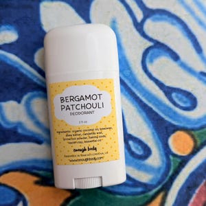 May include: A white plastic deodorant stick with a yellow label featuring white polka dots. The label text reads Bergamot Patchouli Deodorant, 2.5 oz, ingredients list, enough body, Handmade in Rancho Cordova, CA, and www.enoughbody.com.