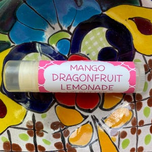 May include: A pink and white lip balm tube with the text "Mango Dragonfruit Lemonade Lip Butter" printed on the label.