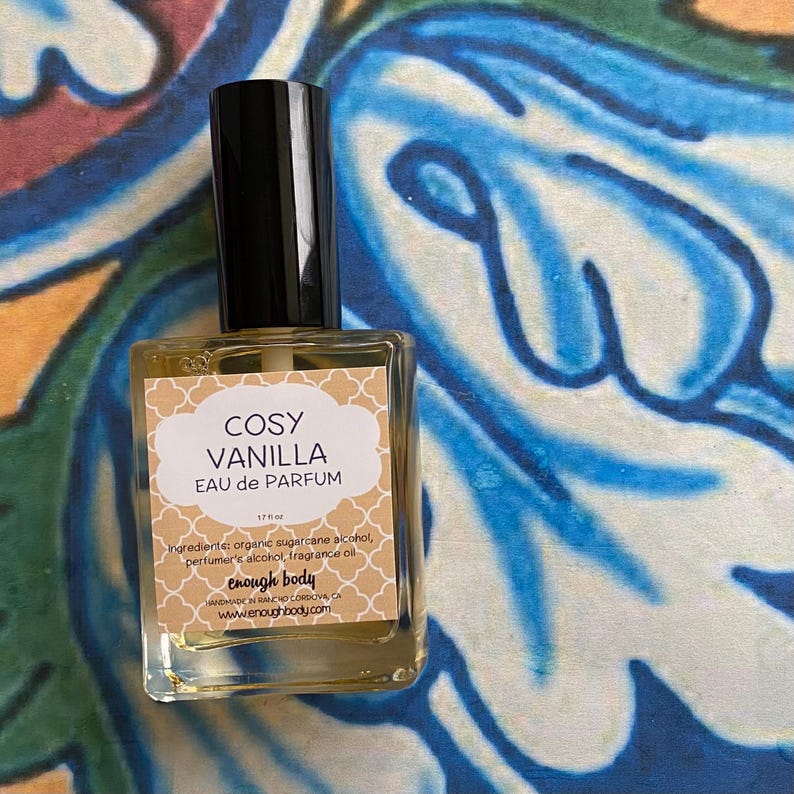 K&ouml;nnte beinhalten: Eine durchsichtige Glasflasche mit schwarzem Verschluss, die eine hellgelbe Fl&uuml;ssigkeit enth&auml;lt. Das Etikett lautet "COSY VANILLA EAU de PARFUM" und listet die Inhaltsstoffe als "organischer Zuckerrohralkohol, Parf&uuml;malkohol, Duft&ouml;l" auf. Das Etikett enth&auml;lt auch den Markennamen "enough body" und die Website-Adresse "www.enoughbody.com".