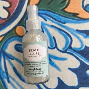 May include: A clear glass bottle with a white spray nozzle and a white label. The label reads "Beach House Sea Salt Hair Mist" and "4 fl oz". The ingredients are listed on the label, including distilled water, aloe vera, sea salt, dead sea salt, argan oil, fragrance oil, and mica. The label also includes the brand name "enough body" and the website address "www.enoughbody.com".