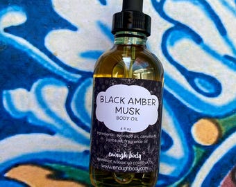 Puede incluir: Una botella de vidrio transparente de Black Amber Musk Body Oil con un cuentagotas negro. La etiqueta es blanca con texto negro y un diseño floral. La botella contiene un líquido de color dorado. El fondo es un diseño abstracto azul y blanco.