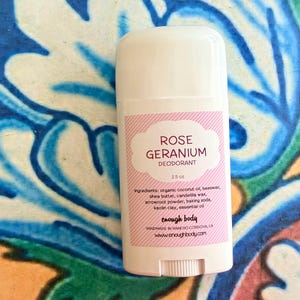 May include: A white plastic deodorant stick container with a pink and white striped label. The label reads Rose Geranium Deodorant 2.5 oz, lists ingredients including organic coconut oil, beeswax, shea butter, and includes the brand name enough body.