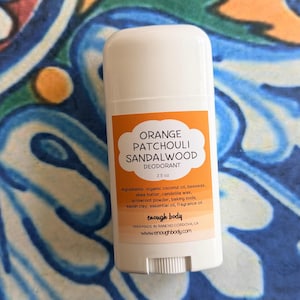 May include: A white plastic deodorant stick container with an orange label. The label text reads Orange Patchouli Sandalwood Deodorant, 2.5 oz, ingredients list, enough body, Handmade in Rancho Cordova, CA, and the website address www.enoughbody.com.