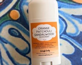 May include: A white plastic deodorant stick container with an orange label. The label text reads Orange Patchouli Sandalwood Deodorant, 2.5 oz, ingredients list, enough body, Handmade in Rancho Cordova, CA, and the website address www.enoughbody.com.