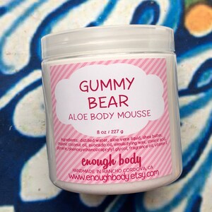 May include: A white plastic jar of "Gummy Bear Aloe Body Mousse" with a pink and white label. The label includes the product name, ingredients, and the brand name "enough body". The jar contains 8 oz of product.