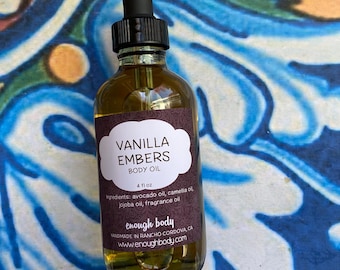 May include: A glass bottle of Vanilla Embers Body Oil with a black dropper. The label displays the text "Vanilla Embers Body Oil" and lists ingredients. The bottle contains 118 ml of oil and is set against a colourful, patterned background.