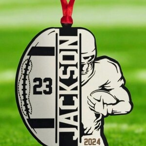 May include: A football-themed ornament featuring the name "JACKSON" and the number "23". The year "2024" is also visible. The design includes a football player graphic and is hung with a red ribbon, suitable for festive decoration.