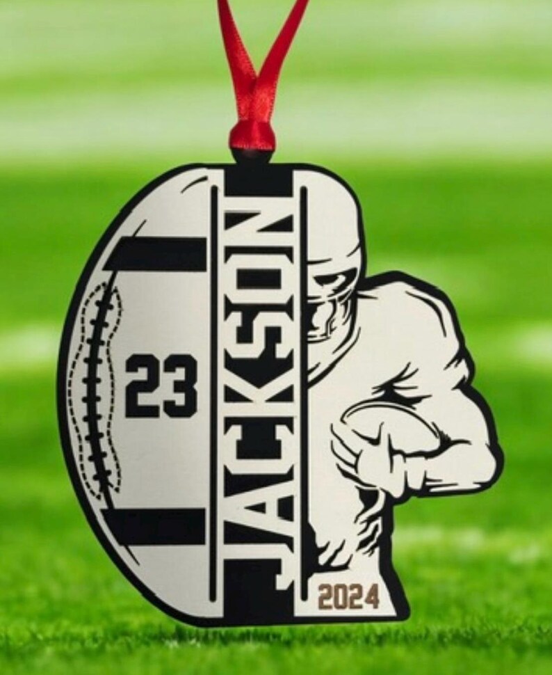 May include: A football-themed ornament featuring the name "JACKSON" and the number "23". The year "2024" is also visible. The design includes a football player graphic and is hung with a red ribbon, suitable for festive decoration.