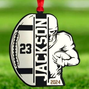 May include: A football-themed hanging ornament with a red ribbon. The design includes a football shape with the number "23" and the name "JACKSON". A football player graphic is on the right, with the year "2024" at the bottom.