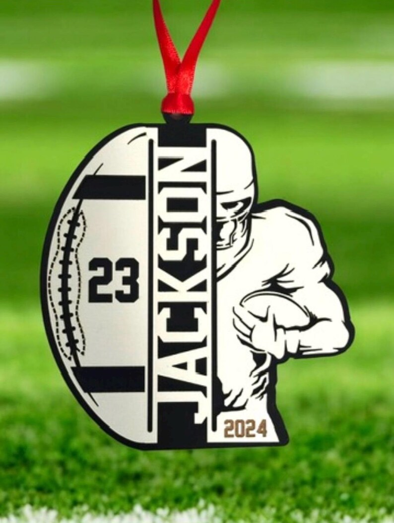 May include: A football-themed hanging ornament with a red ribbon. The design includes a football shape with the number "23" and the name "JACKSON". A football player graphic is on the right, with the year "2024" at the bottom.