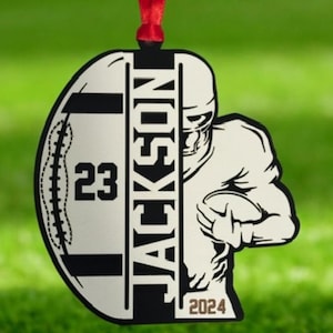 May include: A football-themed ornament with a red ribbon. The ornament features a football shape with the name "JACKSON" vertically, the number "23", and a football player graphic holding a ball. The year "2024" is also displayed.