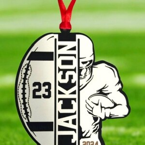 May include: A football-shaped ornament featuring the name "JACKSON" in black. The number "23" is displayed on the left, with a football player graphic on the right. The year "2024" is at the bottom. A red ribbon is attached for hanging.