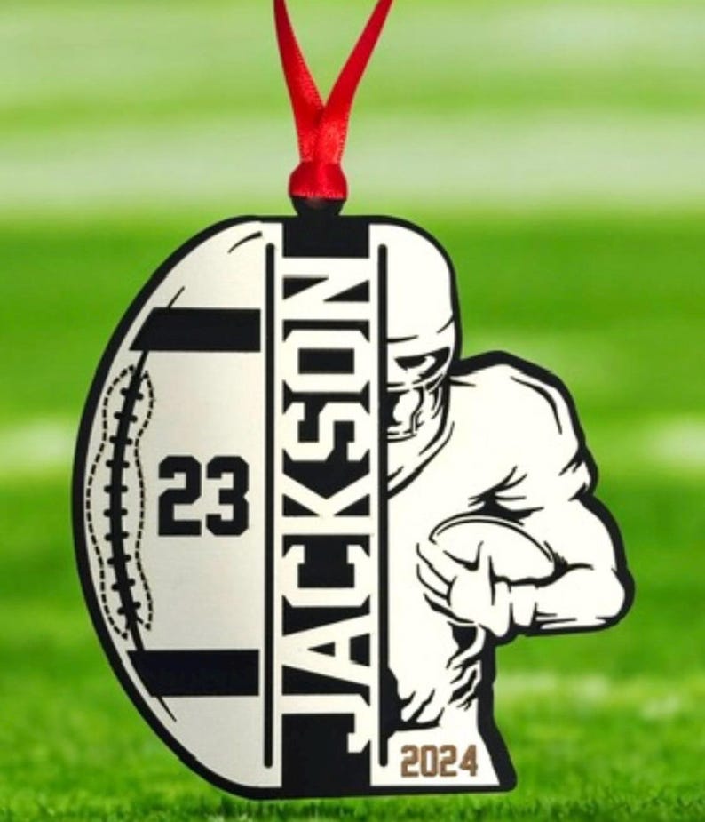 May include: A football-shaped ornament featuring the name "JACKSON" in black. The number "23" is displayed on the left, with a football player graphic on the right. The year "2024" is at the bottom. A red ribbon is attached for hanging.