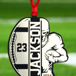 May include: A football-shaped ornament with a red ribbon. The ornament features a football player graphic and the name "JACKSON". The number "23" and the year "2024" are also displayed. Ideal for sports fans.
