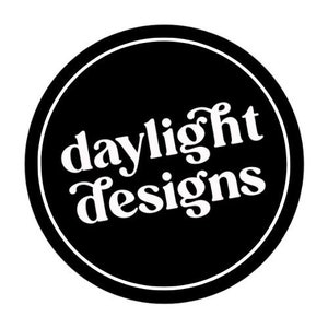 May include: A circular logo featuring a black background and a white border. The words "daylight designs" are written in a white, stylized font within the circle.