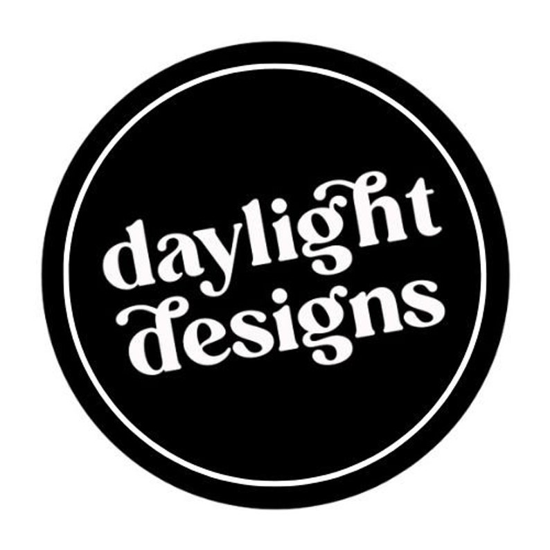 May include: A circular logo featuring a black background and a white border. The words "daylight designs" are written in a white, stylized font within the circle.