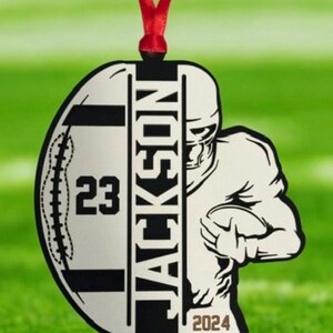 May include: A football-shaped ornament with a red ribbon. The ornament features a football player graphic, the name "JACKSON", the number "23", and the year "2024". The ornament is white and black.