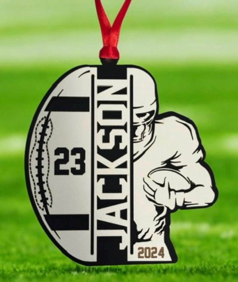 May include: A football-shaped ornament with a red ribbon. The ornament features a football player graphic, the name "JACKSON", the number "23", and the year "2024". The ornament is white and black.