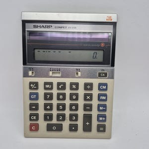 May include: A vintage Sharp Compet VX-2126 calculator with a silver and beige body. The calculator features a 12-digit display and numerous function keys, including square root, percentage, and memory functions. The text "SOLAR CELL CALCULATOR" is visible.