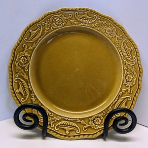 May include: A brown ceramic plate with a decorative raised design around the edge. The design features stylized leaves and flowers. The plate is on a black metal stand.