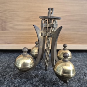 May include: Antique brass clock pendulum with four spherical weights. The pendulum has a central twisted rod and a gear mechanism at the top. The weights are approximately 1.5 inches in diameter. The overall design is ornate and detailed.