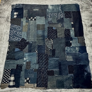May include: A rectangular textile quilt constructed from various shades of blue fabric patches. The design features a mix of solid indigo, polka dot, and wave-patterned squares stitched together with visible white thread in a grid-like arrangement.