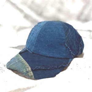 May include: A baseball cap constructed from multiple panels of blue denim fabric. The design features visible frayed edges along the seams and a contrasting lighter blue denim patch on the left side of the curved brim, creating a textured look.