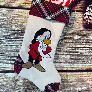 May include: A Christmas stocking with a cream-colored body and a red, black, and white plaid cuff and accents. The stocking features an embroidered image of a cartoon character in a red coat and black pants.