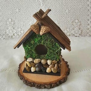 Może przedstawiać: Mały domek dla ptaków wykonany z drewna i ozdobiony mchem, kamieniami i szyszkami. Domek stoi na drewnianym plastrze.