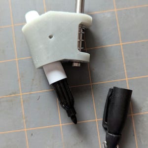 May include: A white 3D printed pen holder with a black marker inserted. A second black marker is lying next to the holder.