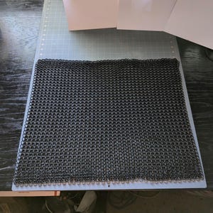 Peut inclure: Une feuille de cotte de mailles noire, tissée à partir d'anneaux métalliques, est posée à plat sur une surface grise.