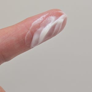Puede incluir: Primer plano de un dedo con una porci&oacute;n de crema blanca. La crema tiene un aspecto liso, ligeramente texturizado, con crestas visibles y un brillo brillante. El fondo es gris claro liso.